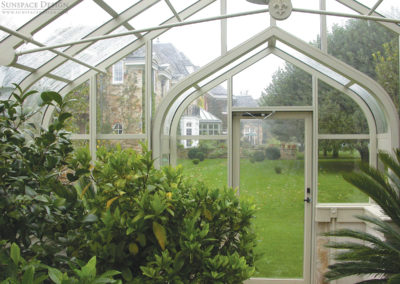 An elegant white aluminum greenhouse with decorative elements and a curved archway adjacent to its glass windows and doors