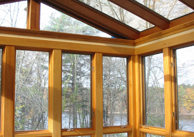 An empty sunroom framed in mahogany and outfitted with large panes of high performance glass reveals views of a nearby pond