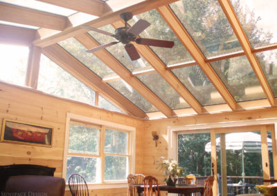 A ceiling fan hangs from a fixture located on the center ridge beam of a sunroom's glass roof