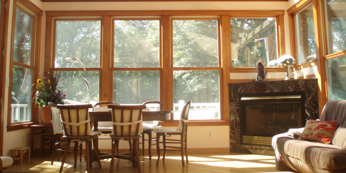 Walls of double hung windows surround a dining table, entertainment area, and corner gas-fired fireplace installation