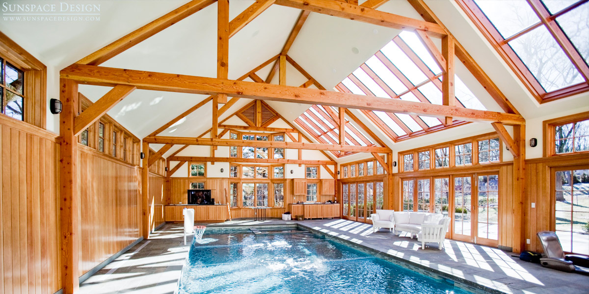 An interior photograph of a indoor swimming pool enclosure featuring rustic wood beams and an expansive glass roof system designed and constructed by Sunspace Design in Newton, Massachusetts