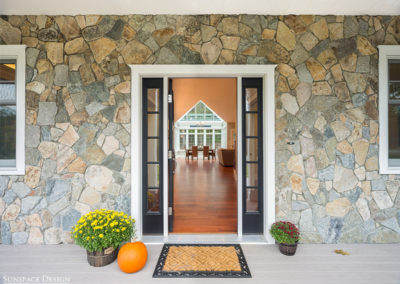 An exterior perspective through the front door of the home, across the hardwood floors of the great room, and toward a brand new glass conservatory