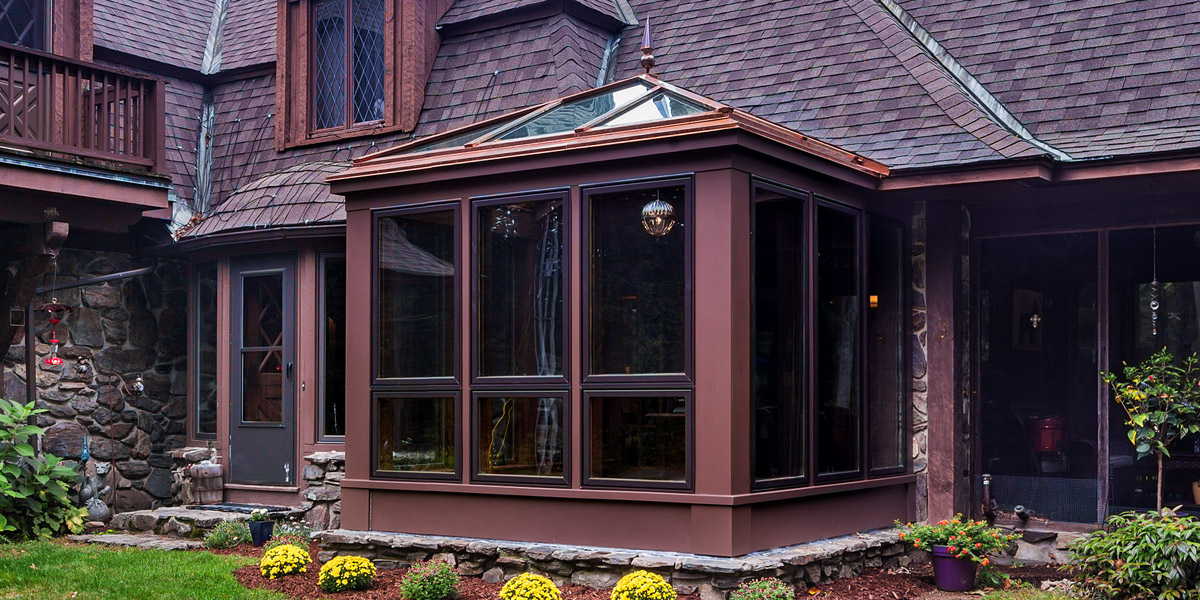 A photo of the exterior of a custom glass conservatory with a hip-style glass roof system, finial, and Marvin casement windows