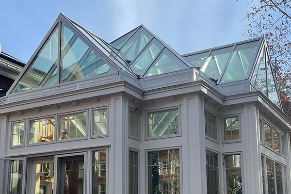 A modern conservatory space with tall windows and angular glass roof components separates the kitchen and living areas of an open concept floor plan